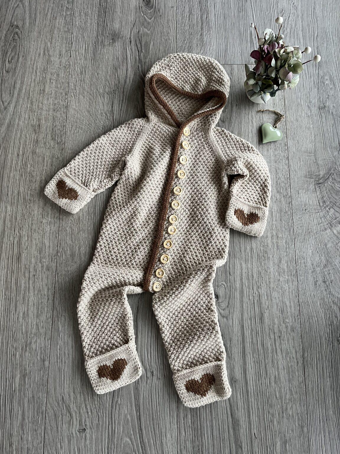 Strikket baby sovedragt med hætte og knapper, dekoreret med hjerte-detaljer, placeret på et træbord ved siden af blomster.