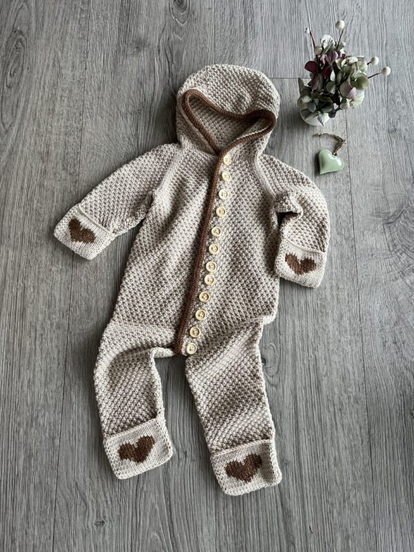 Strikket baby sovedragt med hætte og knapper, dekoreret med hjerte-detaljer, placeret på et træbord ved siden af blomster.