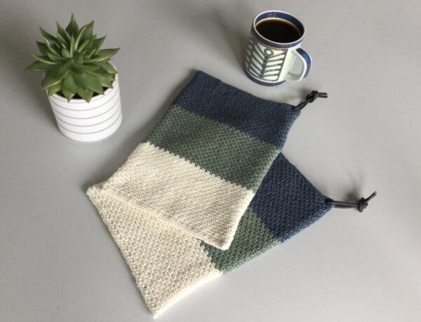 Hæklede grydelapper i blå, grøn, hvid ved siden af en kop kaffe og en grøn plante i potte på gråt bord.