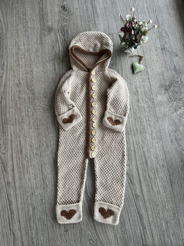 Beige strikkedragt til baby med hætte og hjerter, liggende på et trægulv ved siden af blomster og et grønt hjerte.