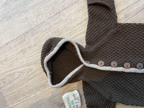Håndstrikket brun babycardigan med stilfulde knapper og hætte ligger på trægulv. Kort tekst ved siden af.