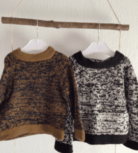 Strikket sweater til børn i forskellige mønstre og farver hænger på en gren mod en hvid væg.