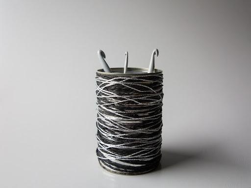 Beholder af metaldåse to hæklenåle i neutral baggrund. Sort og hvidt garn viklet rundt om beholder.