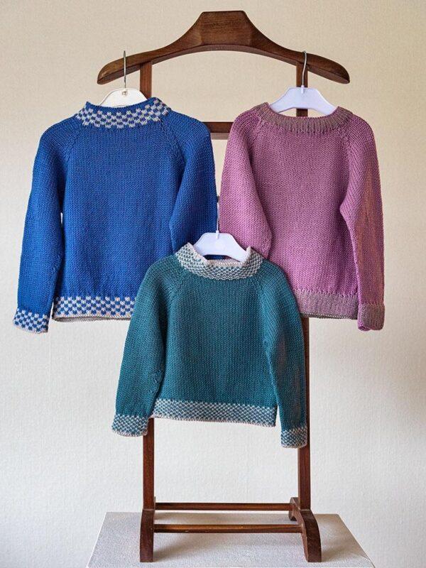 Blå sweater. Tre farverige, strikkede trøjer i blå, grøn og lilla nuancer hænger på bøjler.
