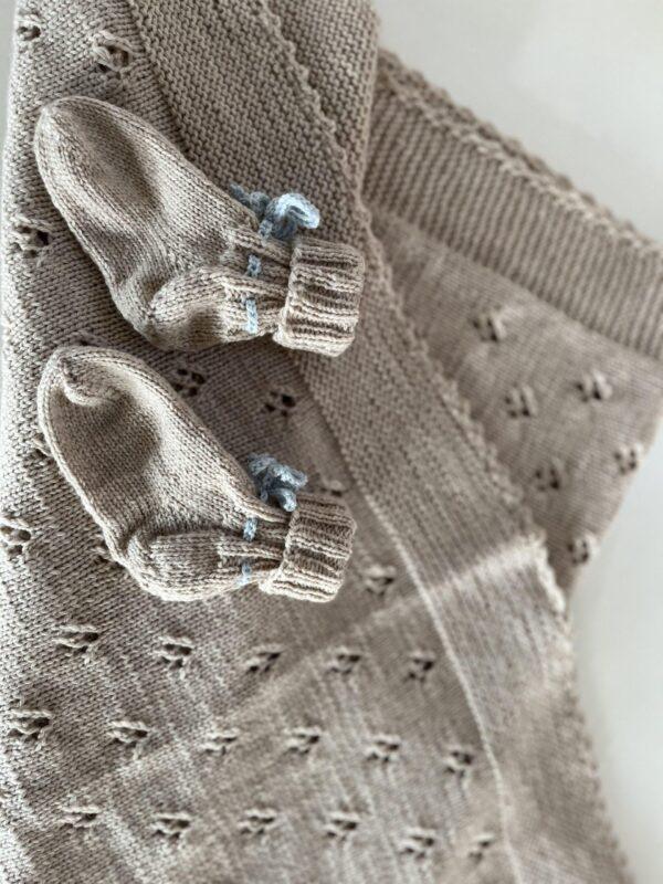 Beige strikket babytæppe med mønster og små matchende sokker på toppen.