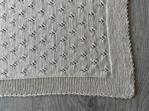 Strikket Beige strikket babytæppe med mønster på trælaminatgulv. med detaljemønster på grå trægulv. Håndlavet design med dekorative åbninger og tekstur.