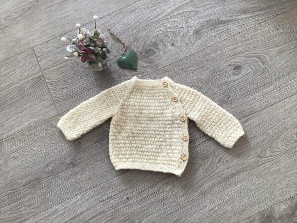 Strikket babytrøje med knapper på en træplade, ved siden af en tørret blomsterbuket og et grønt hjerte.