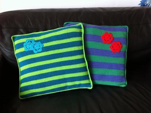 To stribede hæklede puder med hæklede blomster på en sort sofa. Farverne er grøn, blå og lilla med blomster i rød og turkis. Gratis opskrift