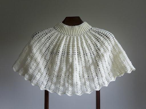 Hvid hæklet cape med flot struktur, i delikat mønster fremvist på en mørk baggrund. Elegant og tidløst design til enhver garderobe.