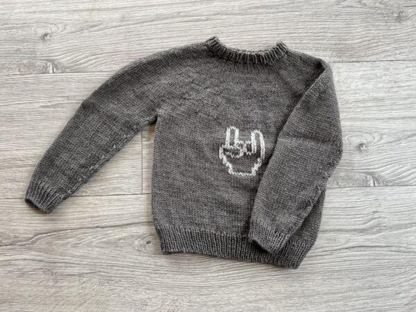 Grå strikket sweater med rock on motiv foran, perfekt til børn, lagt ud på en lys trægulvbaggrund.