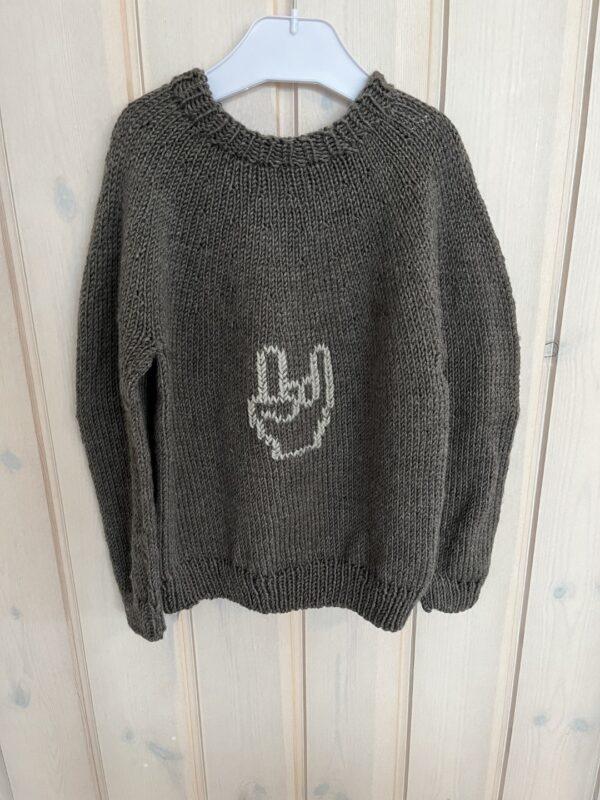 Grå sweater med hvidt håndtegn hænger på trævæg, strikket i kraftigt garn.