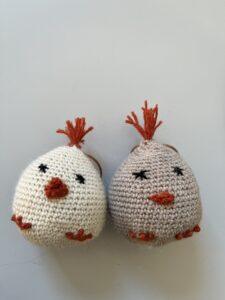 Crocheted Easter chicks with red details on a light surface, one in beige and one in white, standing side by side.