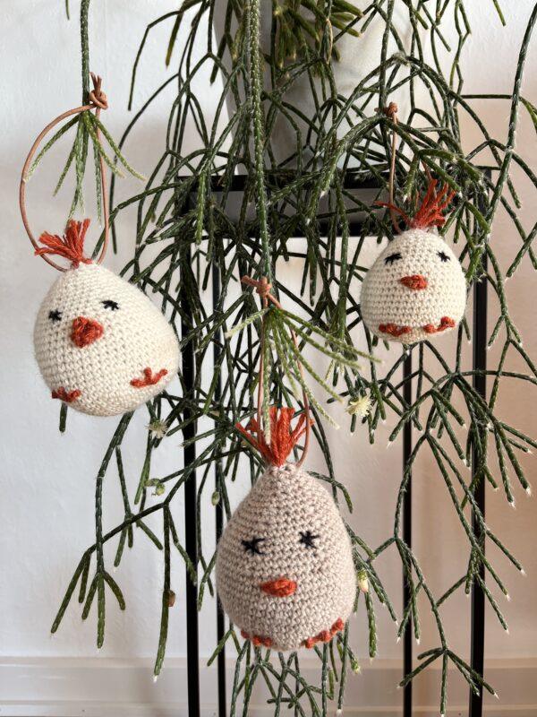 Hæklede påskekyllinger. Tre hæklede fuglepynt med orange næb og totter, hængende fra en grøn plante i minimalistiske omgivelser.