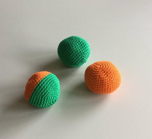 Hacky sack - Tre strikkede bolde i grønne og orange farver liggende på en lys baggrund.