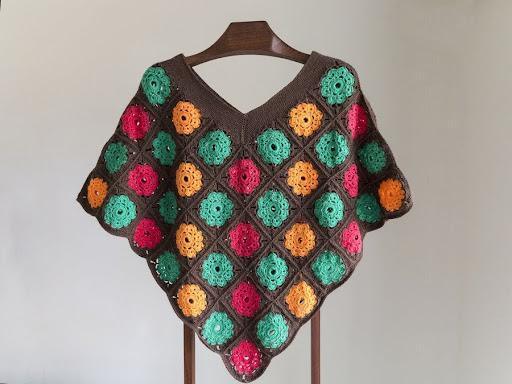 Poncho dame - Håndlavet farverig poncho med blomstermotiv i orange, grøn og rød på brun baggrund, udstillet på en bøjle.
