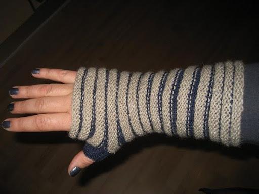Opskrift på pulsvarmer med tommelfinger. Hånd iført en strikket, stribet fingerløs handske i beige og blå nuancer, der strækker sig over håndleddet.