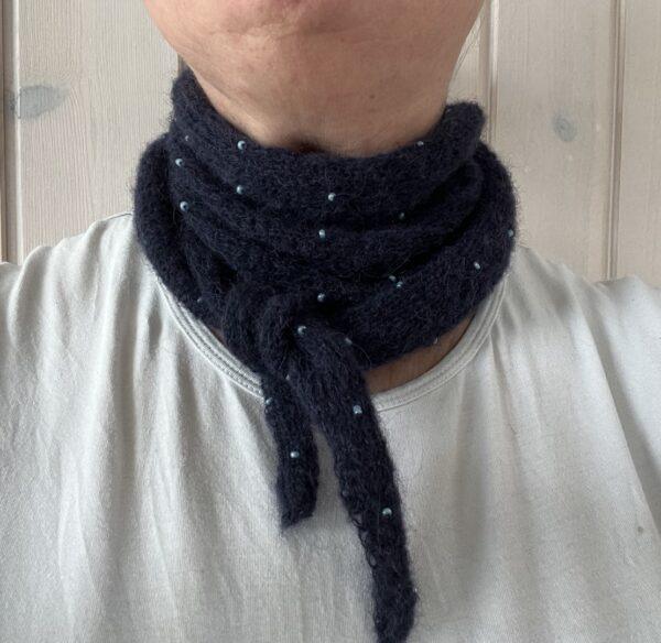 Strikket tørklæde med perler. En person iført et mørkt tørklæde med små blå perler, viklet om halsen over en hvid skjorte.