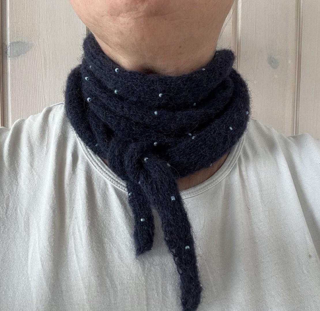 Strikket tørklæde med perler. En person iført et mørkt tørklæde med små blå perler, viklet om halsen over en hvid skjorte.