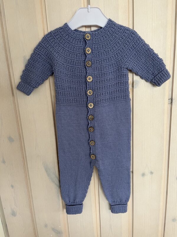 Strikket heldragt til baby. En strikket blå babyjumpsuit med en tekstureret top, knaplukning foran og elastiske manchetter, vist på en bøjle mod træpaneler.