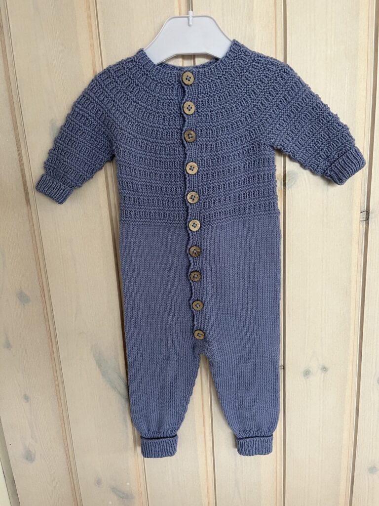 Strikket heldragt til baby. En strikket blå babyjumpsuit med en tekstureret top, knaplukning foran og elastiske manchetter, vist på en bøjle mod træpaneler.