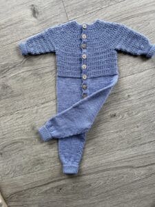Strikket heldragt til baby. En strikket blå babyheldragt med tekstureret overdel, træknapper og lange ærmer lagt på en træoverflade.