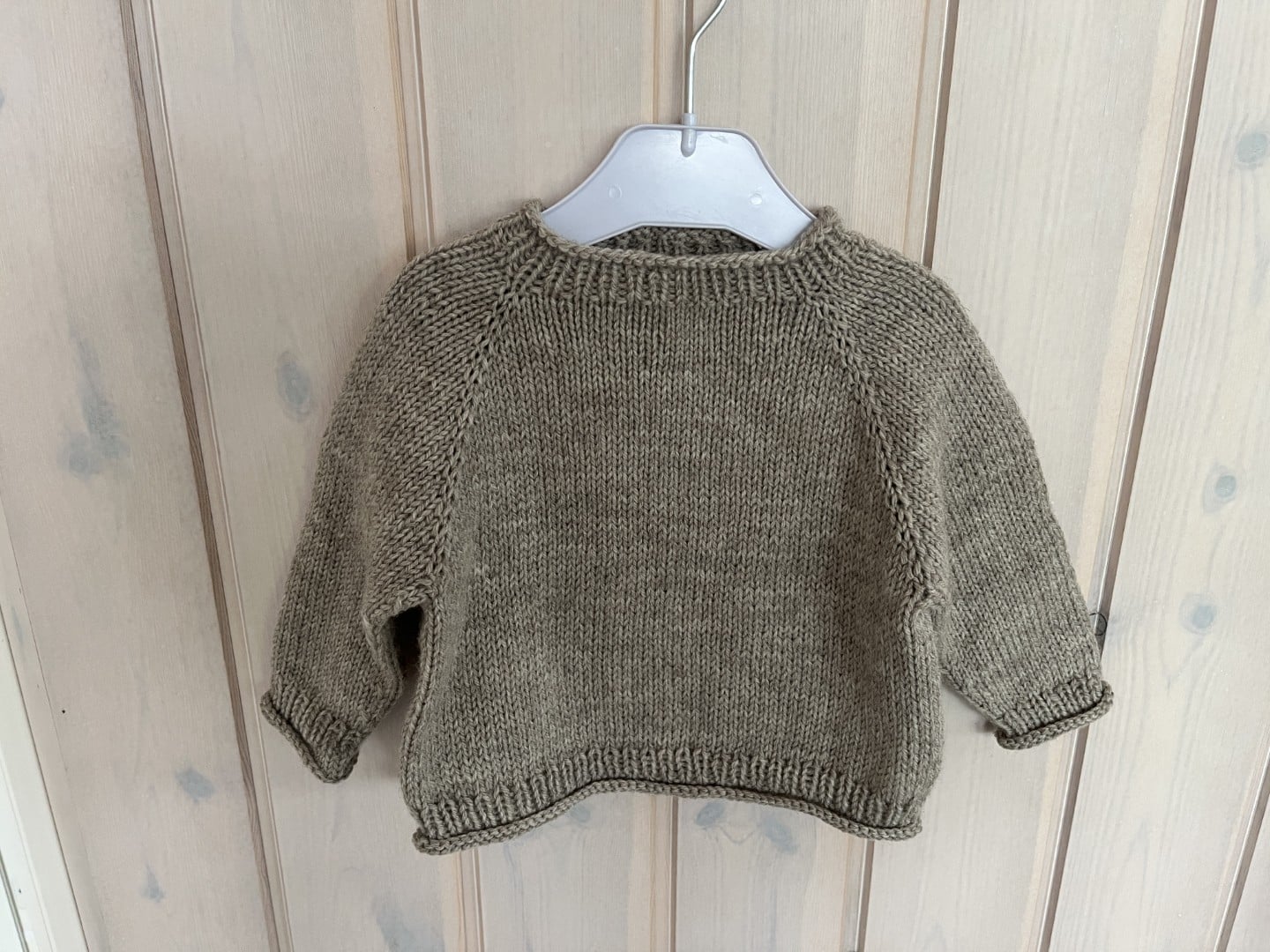 Udendørstrøje / outdoor sweater til baby og barn. En hyggelig, håndlavet brun striktrøje, der hænger på en hvid plastikbøjle mod en træbaggrund.