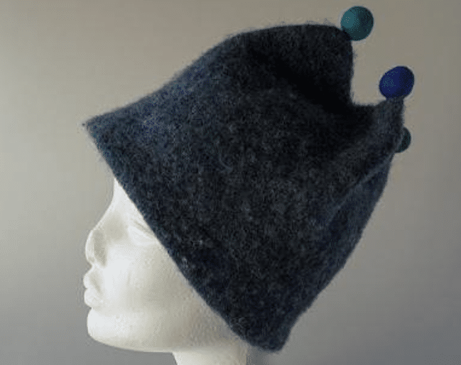 Strikket Saunahue. En blå filthat med en unik form med to farverige pomponer, vist på en hvid buste mod en neutral baggrund.